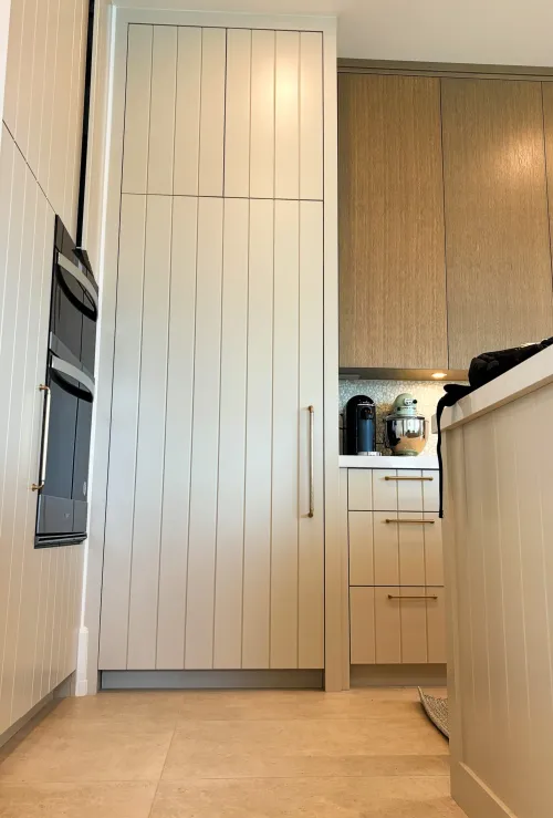 Pool house detail — floor-to-ceiling painted cabinetry tower with vertical-groove panels, wall oven, and oak cabinetry beyond
